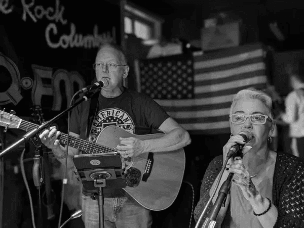 Lela and Dennis Whalen performing with guitar and microphones in front of a Rock Columbus banner